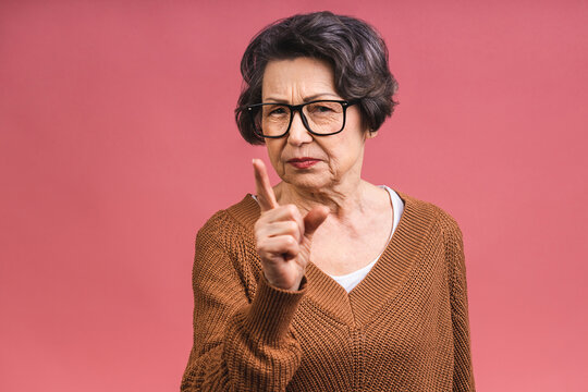 Portrait of angry grey haired old strict senior woman wearing glasses pointing up threatening with finger. Grandmother isolated over pink background.