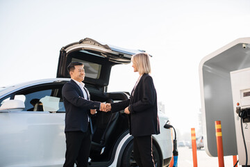 Female customer of dealership shaking hands with competent consultant while buying new auto. Happy caucasian woman and asian man standing together near white luxury electric car outdoors.