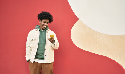 Smiling African American gen z teen standing at color wall using mobile phone. Happy cool ethnic stylish hipster guy model holding cell phone, checking apps on mobile device, looking at cellphone. - obrazy, fototapety, plakaty
