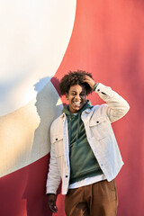 Happy cheerful African American teen guy laughing on red wall lit with sunlight. Smiling cool ethnic gen z teenager student model standing looking at camera, authentic candid shot outdoors. Vertical - obrazy, fototapety, plakaty