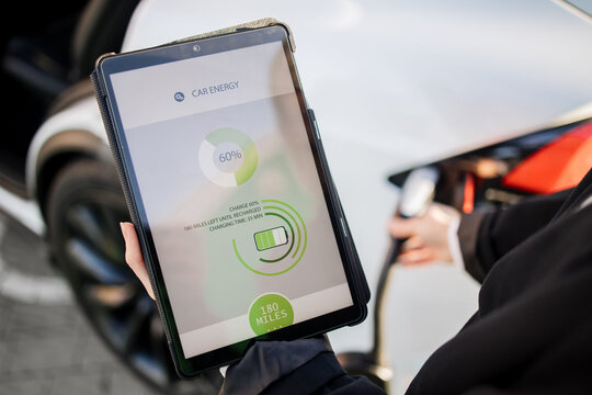 Close up of businesswoman in suit using digital tablet during electric automobile charging session outdoors. Focus on gadget screen with modern app for car.