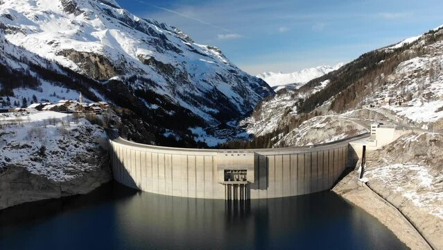 Flyover of hydroeletric dam in French alps in winter snow, across dramatic valley scenery and quaint traditional village