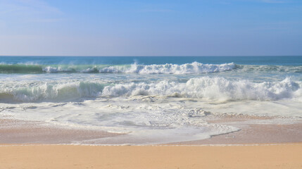Waves and foam in the Atlantic Ocean under a blue sky on a sandy beach on a winter day near Lisbon,...