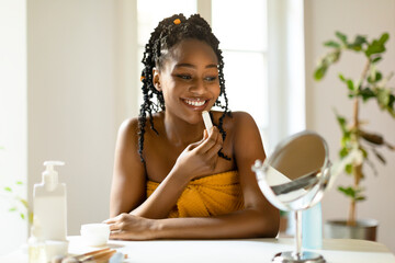 Happy beautiful black woman using moisturizing lipbalm, lady sitting at vanity table, looking at small mirror
