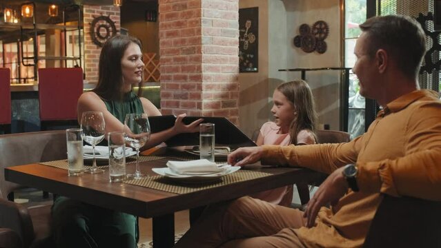 Medium shot of parents and little daughter sitting at table in restaurant and choosing dishes in menu together