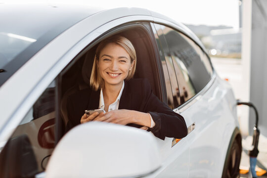 Attractive caucasian woman with modern smartphone in hands sitting inside luxury car while recharging battery on auto station. Business lady in stylish suit working online during trip break.