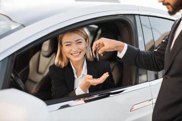 Charming caucasian woman in formal wear siding inside her luxury new car and receiving keys from male dealer. Successful business lady making expensive purchase.