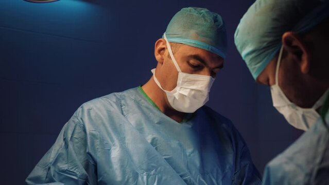 Doctor's face in a medical uniform and a mask makes an operation in the operating room. Doctor's hands takes off medical gloves after surgery. The surgeon has finished his work.