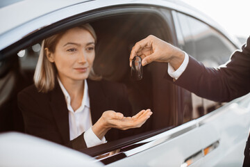 Close up of male seller giving keys from white modern car to happy female owner in formal wear. Charming young woman with blond hair sitting inside her luxury new auto.