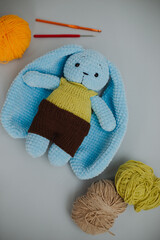 blue knitted rabbit with big ears in a yellow vest and brown pants with three skeins of yarn, brown, yellow, green and knitting needles