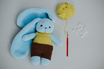 blue knitted rabbit with big ears in a yellow vest and brown pants with a spool of thread and a knitting needle