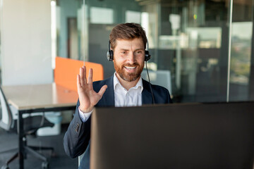 Happy businessman in headset working on computer and waving his hand to webcamera, having video call in office interior