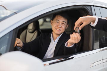 Portrait of pleased asian man in black suit receiving car keys from male dealership seller. Happy male in formal attire buying news luxury vehicle. Concept of successful business and consumerism.