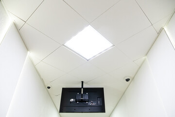 A square lamp and an advertising monitor on a white office ceiling. Round video surveillance lamps in the bank. Interior design in the office. A fluorescent lamp on a modern suspended ceiling.