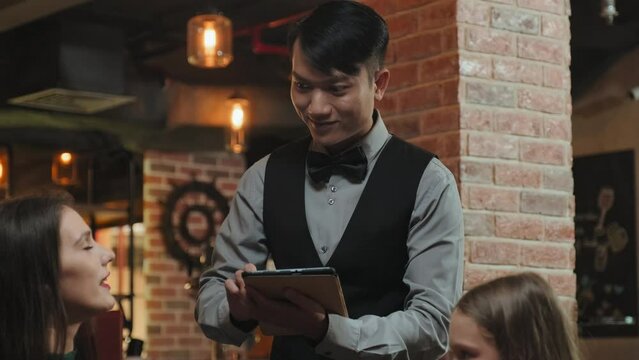 Tilt up shot of woman and her little daughter talking to Asian waiter as he taking order with digital tablet in restaurant
