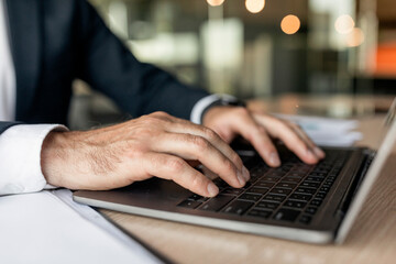 Closeup businessman using laptop, typing on keyboard, writing email, sitting at desk in office, selective focus