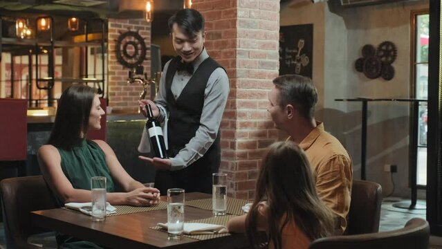 Medium wide shot of waiter in classic uniform bringing bottle to table and recommending wine pairing while serving family at restaurant