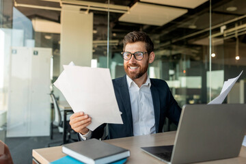 Happy male entrepreneur reading business papers and smiling, checking company growth rates, sitting at desk in office