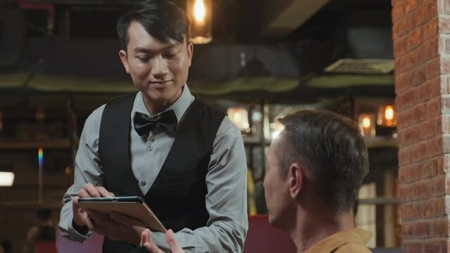 Medium shot of Asian waiter in elegant uniform using app on digital tablet while taking order from man in restaurant