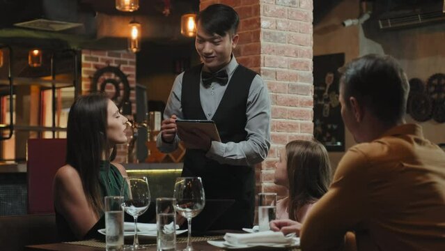 Family speaking with Asian waiter as he taking order with digital tablet in restaurant