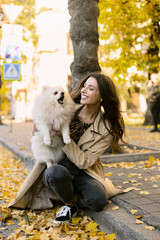 Attractive girl is holding a white dog, the dog is barking, the girl is looking at him. A walk outdoors with a pet.