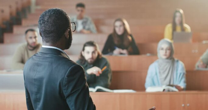 Rear of African American male professor standing in front of students in college and talking to them while having lection. High school. Back view. Man teacher explaining subject at lesson. Close up.