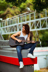 A young, handsome man with long hair and a black guitar, on the streets of the city, in a white T-shirt and jeans