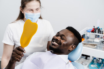 African man looking on teeth in the mirror at the dentist. Happy african american patient of...