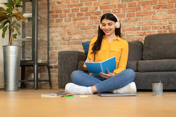 Positive spanish teen lady taking notes in notepad, writing in diary and listening music in...