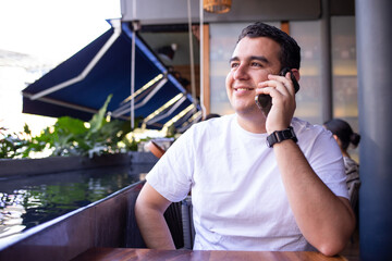 Hombre Latino blanco moreno con una playera blanca en un restaurante tomando una llamada con su...