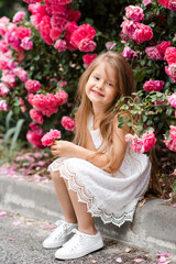 Smiling cute child girl 5-6 year old holding pink rose flower sitting over blooming bushes at nature background outdoor. Childhood. Spring season.