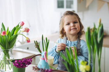 Cute little girl in a pretty blue dress doing home gardening in the kitchen, taking care about flowers and plants. Domestic life, cozy atmosphere, family time, kid's development, hobby, leisure