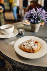 Baked pastry dusted with icing sugar on white plate on top of modern cafe table with purple plant in plant pot and cup of tea in background