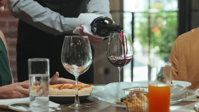 Cropped shot of waiter holding bottle with cloth napkin and pouring red wine into glasses while serving guests in restaurant