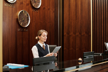 Young blond female receptionist in uniform looking at tablet screen while standing by counter in...