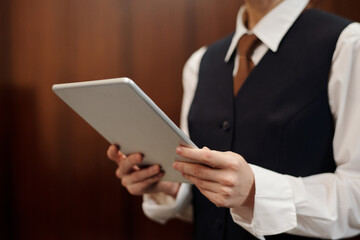 Close-up of young female staff of hotel in uniform using tablet by workplace while looking through...