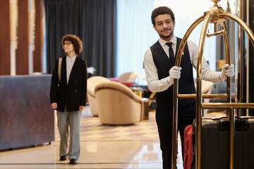 Young bellboy in suit pushing cart with many suitcases of hotel guests while moving along lounge...