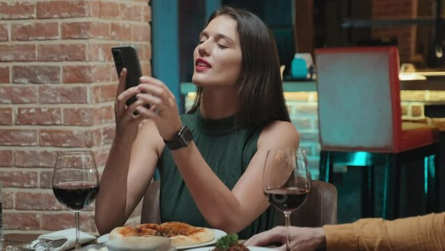 Medium shot of beautiful woman taking pictures of food with smartphone while sitting at table in restaurant