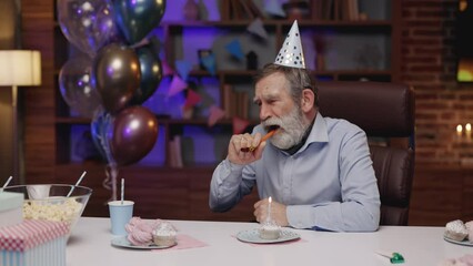 Depressed Mature Man in Party Hat Sitting at Table at Home and Looking Down at Cupcake with Burning Candle, Feeling Unhappy on His Birthday. Loneliness People Concept