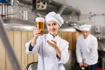 Positive woman brewer in white coat holding beer glass and making presenting gesture.