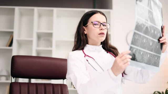 Portrait of concentrated young woman doctor therapist with glasses thinking about diagnosis while looking at results patient MRI or CT scan procedure and looking at camera at light hospital office
