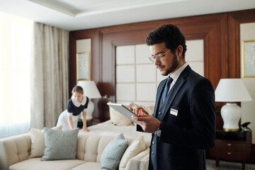 Young elegant entrepreneur in formalwear using tablet while standing in front of camera in hotel room with chambermaid on background - obrazy, fototapety, plakaty