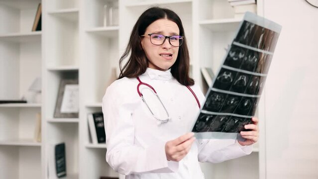 Portrait of doubtful young woman doctor therapist with glasses looking at results patient MRI or CT scan procedure unsure about disease diagnosis and shrugs looking at camera at light hospital 