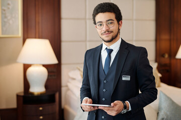 Young confident head manager of luxurious hotel looking at camera while standing against lamp and comfortable double bed in room - obrazy, fototapety, plakaty