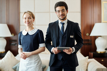 Young successful staff of luxurious five star hotel standing against couch with cushions and two lamps in room prepared for new guests - obrazy, fototapety, plakaty