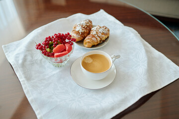 Fresh berry desert, two croissants and cup of coffee on white napkin and plate standing on small brown table in room of luxurious hotel