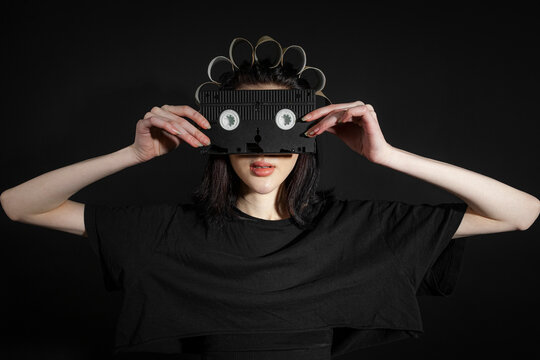 Portrait of a poor, skinny girl in black, rumpled clothes and a strange hat that made of toilet paper tubes holding a vhc cassette imitating glasses, isolated on a black background.