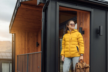 Young female in yellow down jacket walks outdoor near modular cabin house