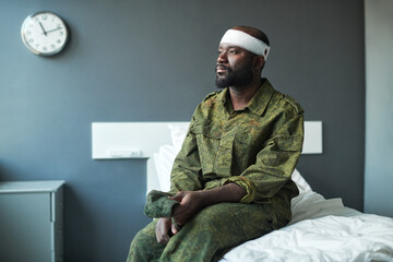 Young African American stressed soldier with bandaged head sitting on bed in hospital ward, looking...