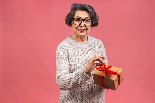 Portrait of senior aged mature beautiful lady woman holding birthday present gift over isolated pink background celebrating achievement with happy smile and winner expression.